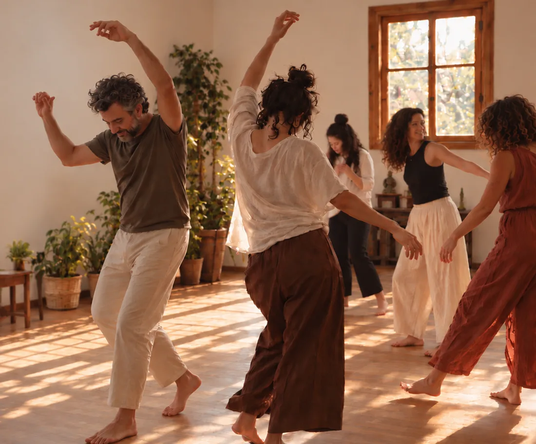 Un grupo de personas descalzas baila en una sala con piso de madera, plantas y luz de tarde.